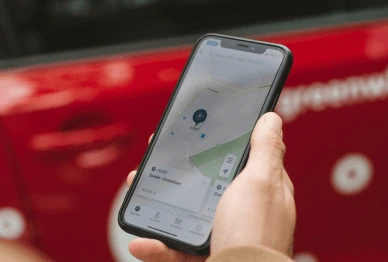 Person holding smartphone with car tracker app in front of red car, showing location on a map.