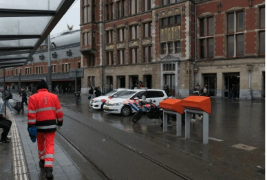 Worker in high-visibility gear walking near parked police cars and orange mailboxes on a wet urban street